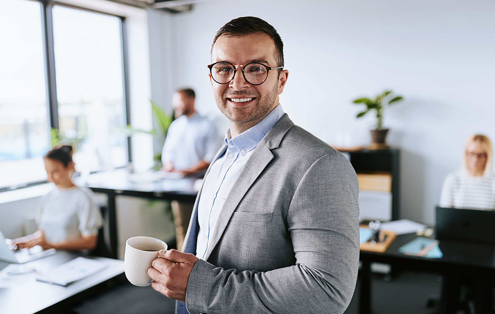 Ein Mann im Anzug h&auml;lt im B&uuml;ro eine Tasse in der Hand und l&auml;chelt in die Kamera.
