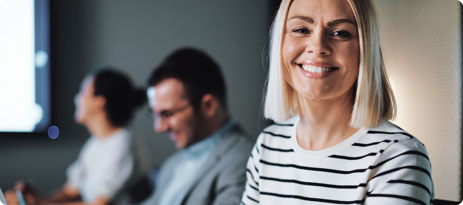 Eine blonde Frau sitzt im B&uuml;ro und l&auml;chelt in die Kamera.